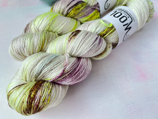 Close up of Two skeins of Image 6 on Luxury 4ply Merino Silk yarn from the Seashells Monthly Yarn club by the hand dyed yarn expert, The Wool Kitchen on a pink and white table. There are also some coloured stitch markers in a small teal pot to the right hand side.