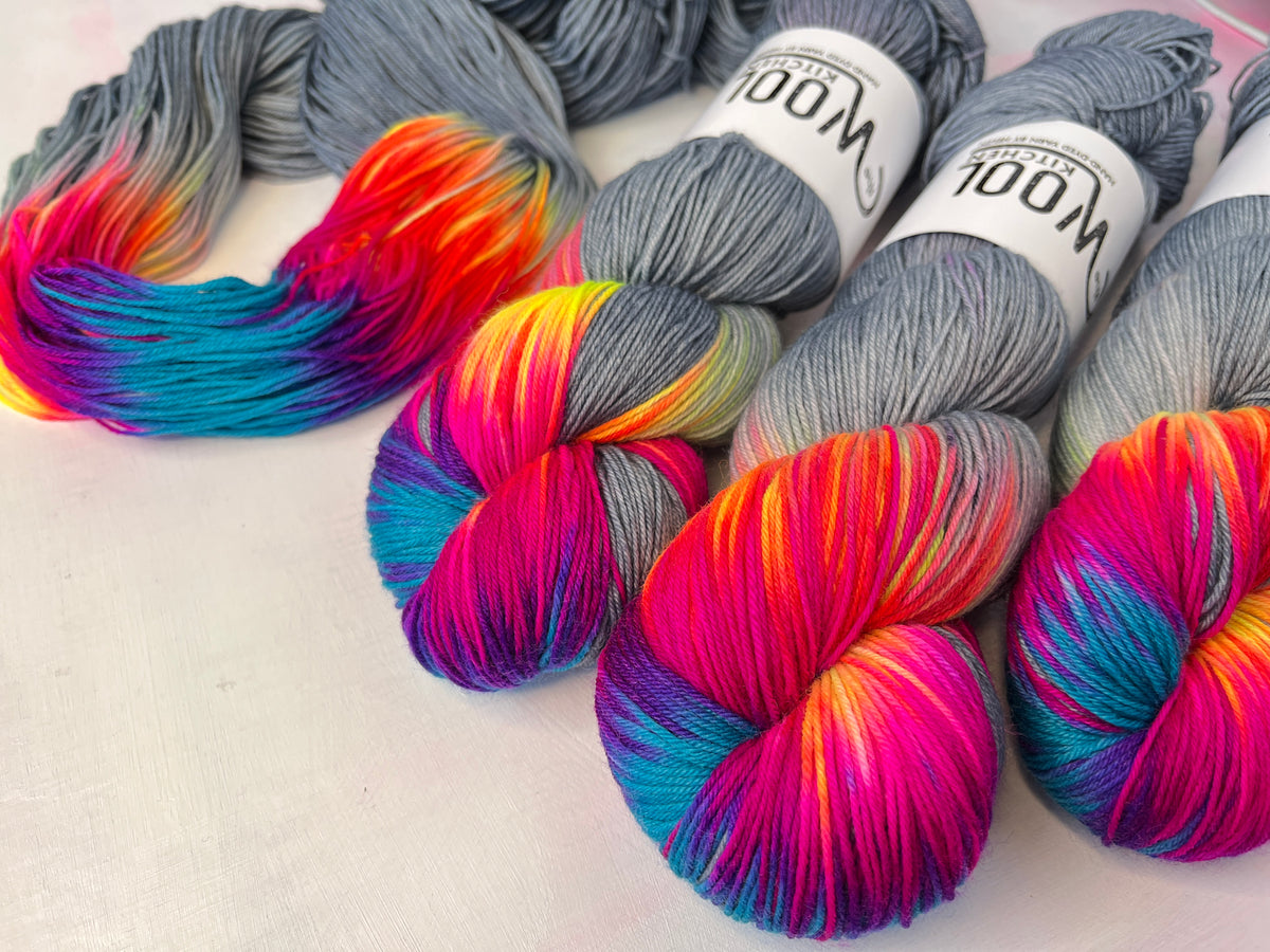 Four skeins of wool yarn with gray bases and vibrant rainbow gradients of pink, purple, blue, orange, and yellow, arranged on a light fabric background. Two skeins have white paper labels reading 'WOOL'.