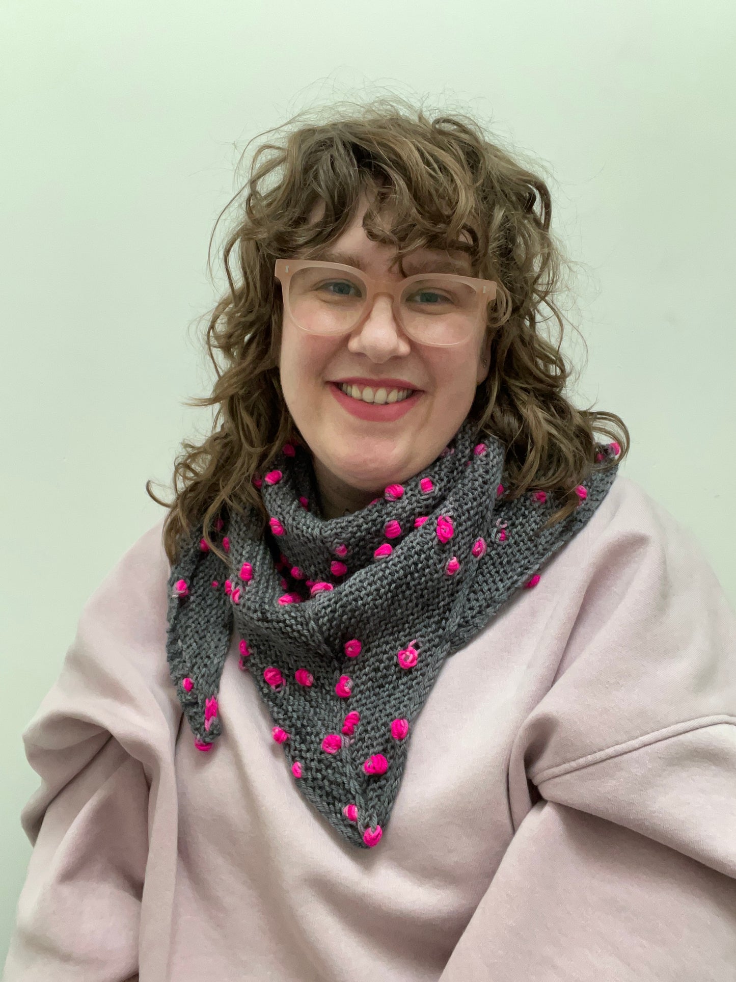 Helen (a white woman with light brown curly hair and glasses) is smiling, looking at the camera wearing a triangular shawl wrapped around her neck. The shawl is dark grey with bright fuchsia pink pops.