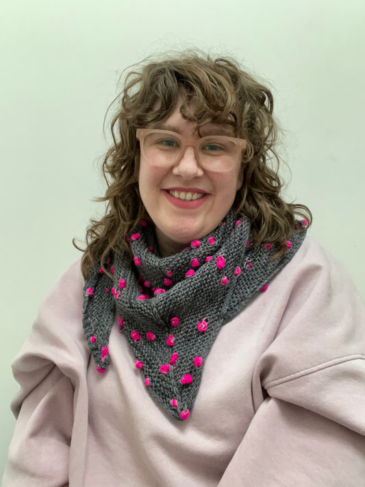 Helen (a white woman with light brown curly hair and glasses) is smiling, looking at the camera wearing a triangular shawl wrapped around her neck. The shawl is dark grey with bright fuchsia pink pops.