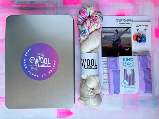 Yarn and knitting accessories including a metal tin, yarn skeins, and packaging on a pink background. The Mother Yarn Gift tin from the hand dyed yarn expert, The Wool Kitchen featuring Champagne Supernova Luxury 4ply Merino Silk yarn.  Perfect for Mothers Day or Easter gifting. 