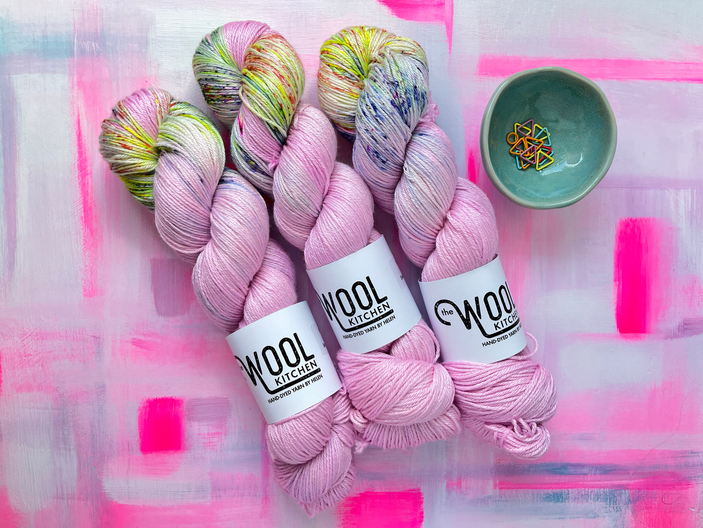 Three skeins of Image 5 on Luxury 4ply Merino Silk yarn from the Seashells Monthly Yarn club by the hand dyed yarn expert, The Wool Kitchen on a brightly painted pink table. There are also some coloured stitch markers in a small teal pot to the right hand side.