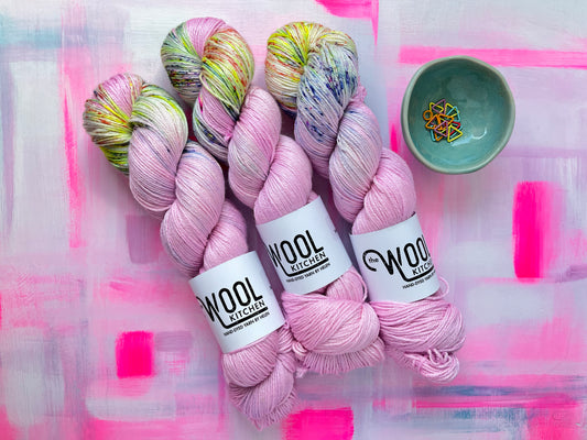Three skeins of Image 5 on Luxury 4ply Merino Silk yarn from the Seashells Monthly Yarn club by the hand dyed yarn expert, The Wool Kitchen on a brightly painted pink table. There are also some coloured stitch markers in a small teal pot to the right hand side.