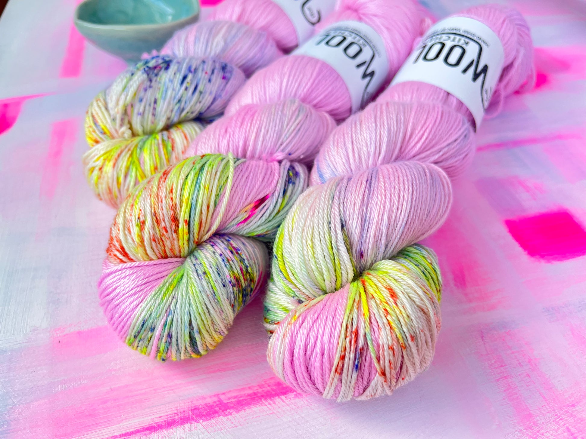 Close up of the zip end of the yarn of Image 5 on Luxury 4ply Merino Silk yarn from the Seashells Monthly Yarn club by the hand dyed yarn expert, The Wool Kitchen on a brightly painted pink table. There are also some coloured stitch markers in a small teal pot to the right hand side.