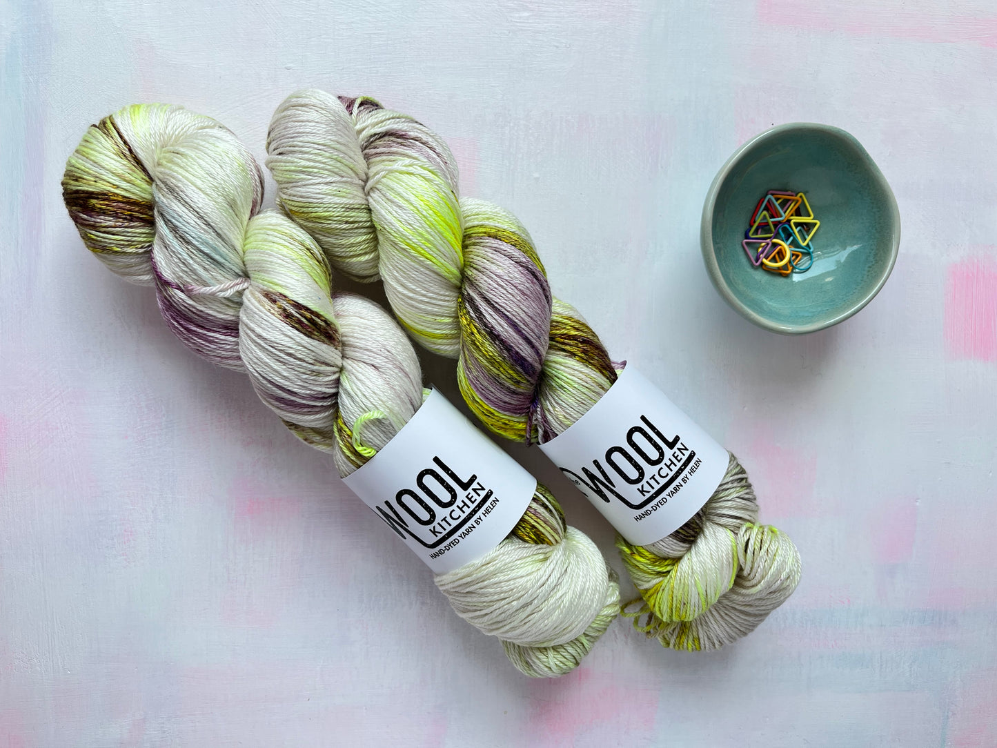 Two skeins of Image 6 on Luxury 4ply Merino Silk yarn from the Seashells Monthly Yarn club by the hand dyed yarn expert, The Wool Kitchen on a pink and white table. There are also some coloured stitch markers in a small teal pot to the right hand side.