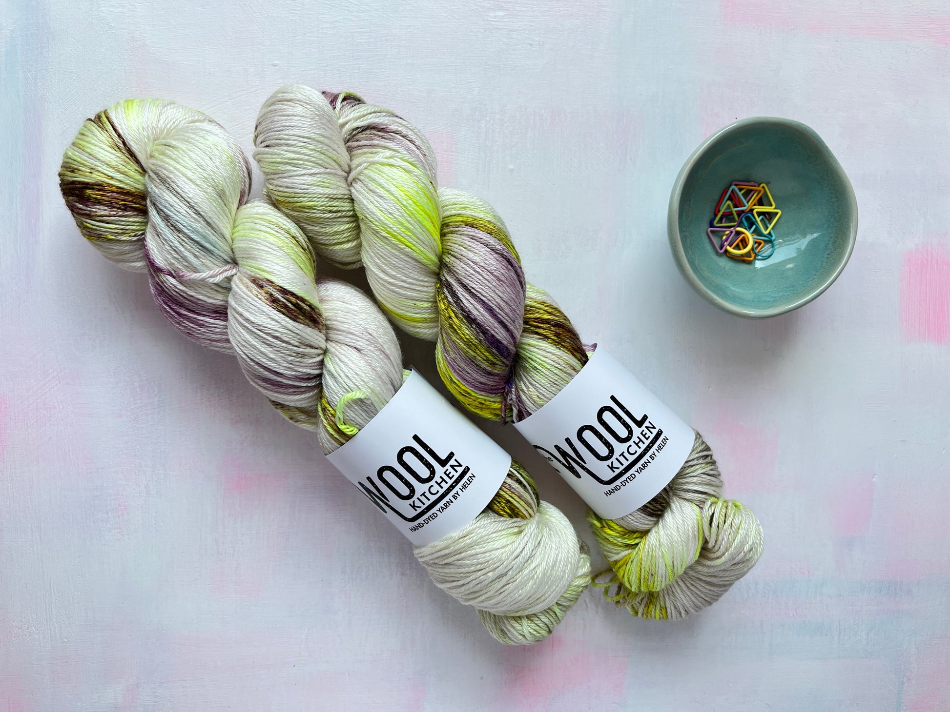 Two skeins of Image 6 on Luxury 4ply Merino Silk yarn from the Seashells Monthly Yarn club by the hand dyed yarn expert, The Wool Kitchen on a pink and white table. There are also some coloured stitch markers in a small teal pot to the right hand side.