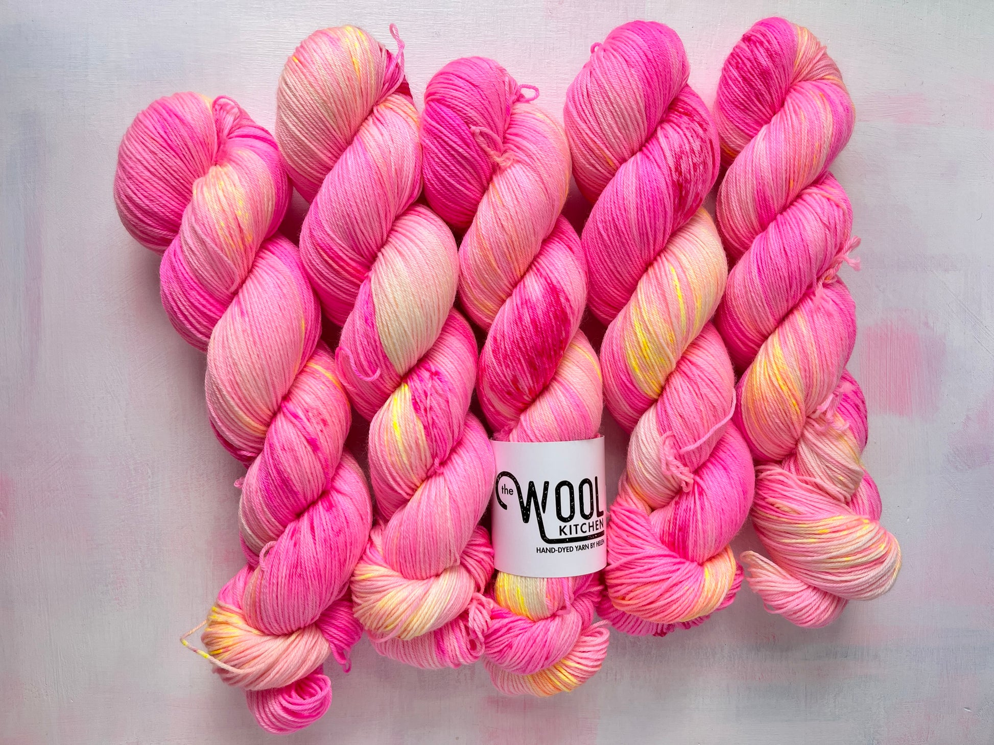 Five skeins of variegated pink, yellow, and white yarn arranged on a light gray background, with a white label reading 'Wool Kitchen' visible near the center.