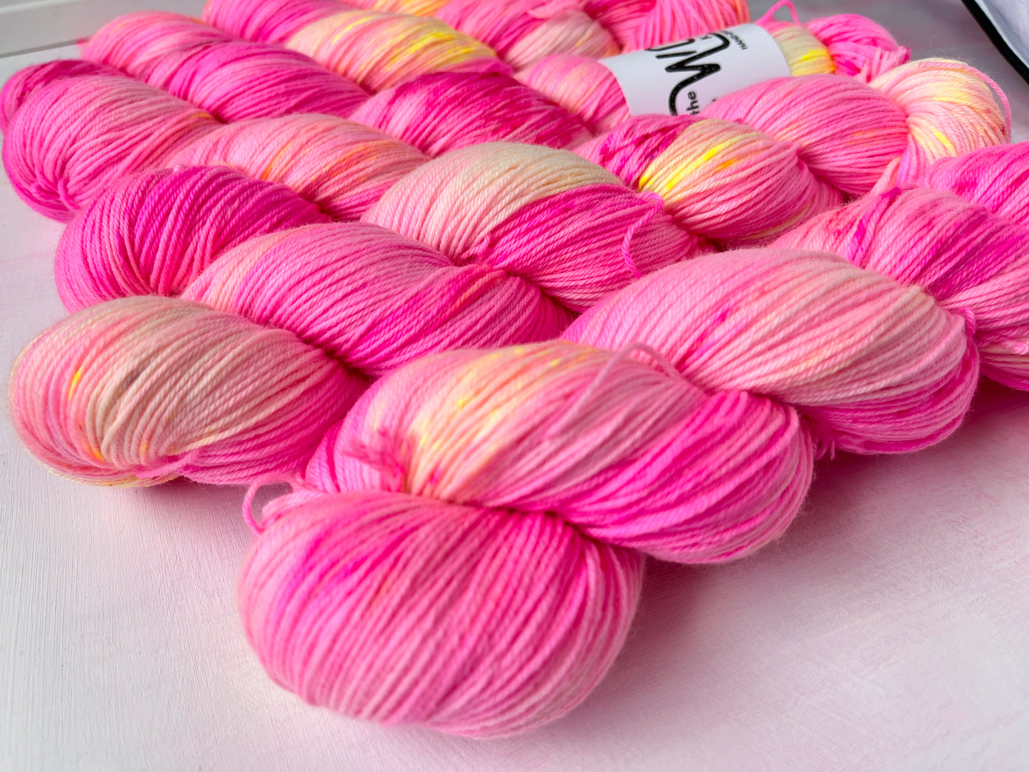 Several skeins of variegated pink yarn arranged on a light surface. The yarn shows alternating bands of bright pink, soft peach, pale cream, and hints of pale yellow.