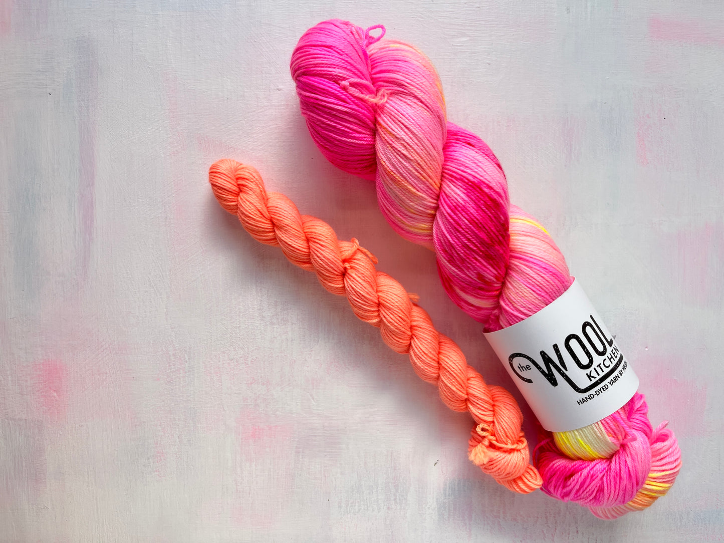 A skein of variegated yarn with a white label reading 'Wool Kitchen' and a matching twisted sample of the yarn, photographed on a light textured background from the July Sock Set for the Floral Fade 3 Fungi Collection by the hand dyed yarn expert. The Wool Kitchen, 