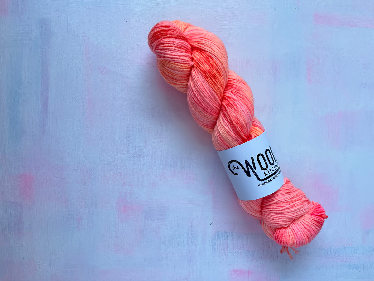 A skein of variegated yarn with a white label reading 'Wool Kitchen' and a matching twisted sample of the yarn, photographed on a light textured background from the August Sock Set for the Floral Fade 3 Fungi Collection by the hand dyed yarn expert. The Wool Kitchen, 