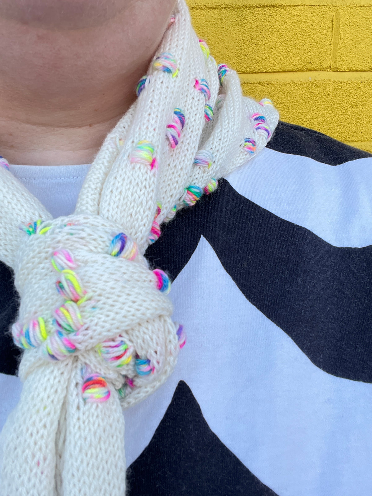 A close-up of a cream-colored knit scarf with colorful rainbow yarn accents, tied around a person's neck over a black and white patterned shirt, with a yellow brick wall in the background. 101 Assign It Bandana Pattern by Helen Reed in Champagne Supernova Zip Yarn on 4ply Sock from the Wool Kitchen.