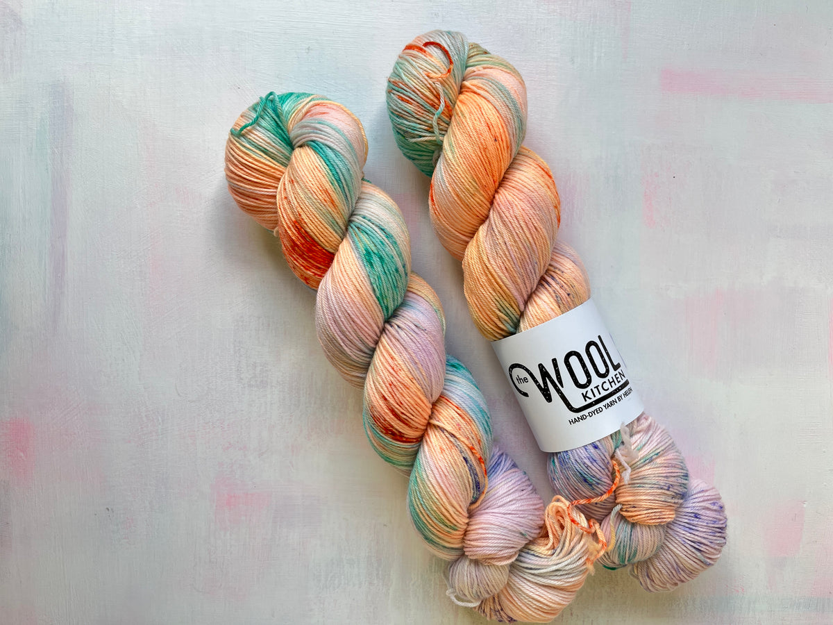 Two hanks of variegated yarn with peach, teal, pink, and lavender tones, laid on a light textured background. One hank has a white label reading WOOL KITCHEN.